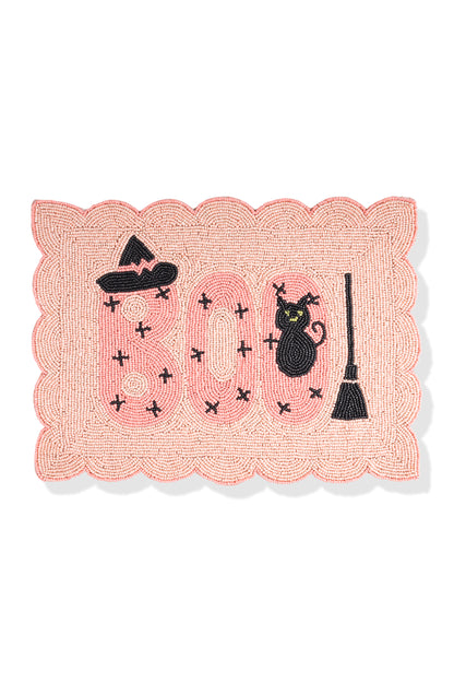 A rectangular woven Halloween mat with a scalloped pink border, featuring the word 'BOO' in pink with black stitched crosses, a black witch hat, a black cat, and a black broomstick on a white background.