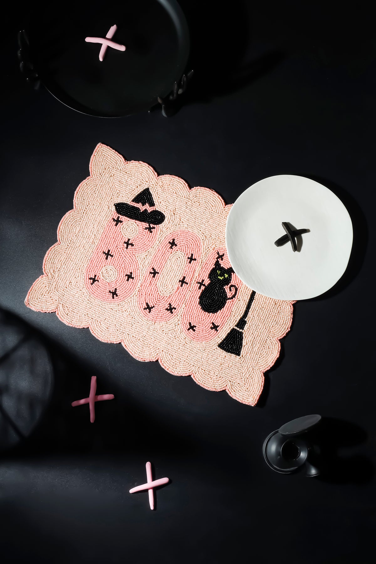 A scalloped pink lace placemat with a Halloween theme, featuring the word 'BOO' in pink, black witch hats, a black cat, and small black stars. It is styled on a dark background with a white plate and pink decorative crosses.