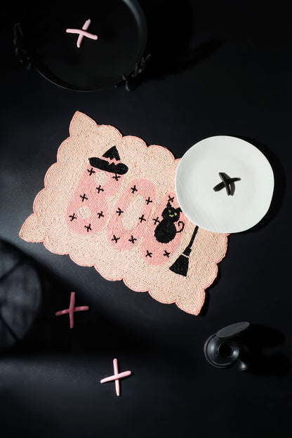A scalloped pink lace placemat with a Halloween theme, featuring the word 'BOO' in pink, black witch hats, a black cat, and small black stars. It is styled on a dark background with a white plate and pink decorative crosses.