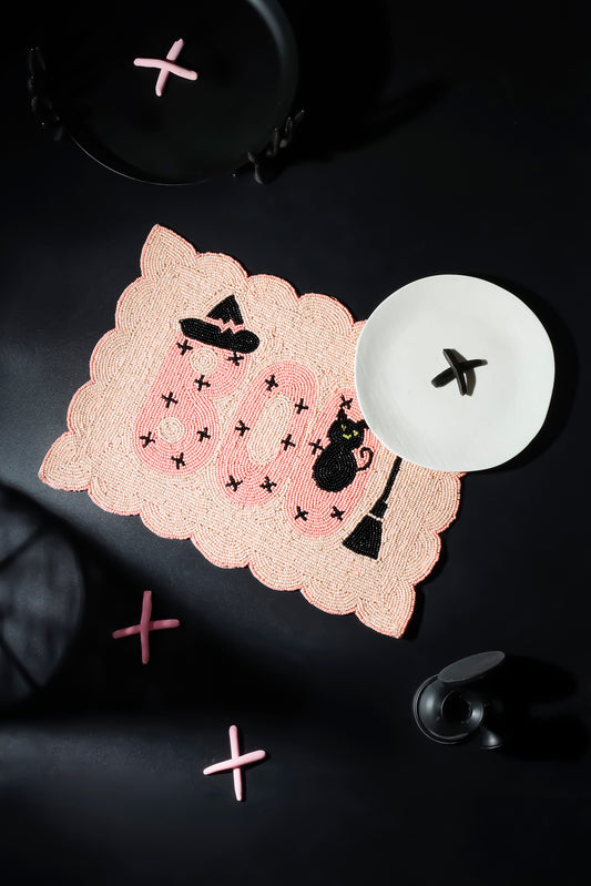 A scalloped pink lace placemat with a Halloween theme, featuring the word 'BOO' in pink, black witch hats, a black cat, and small black stars. It is styled on a dark background with a white plate and pink decorative crosses.