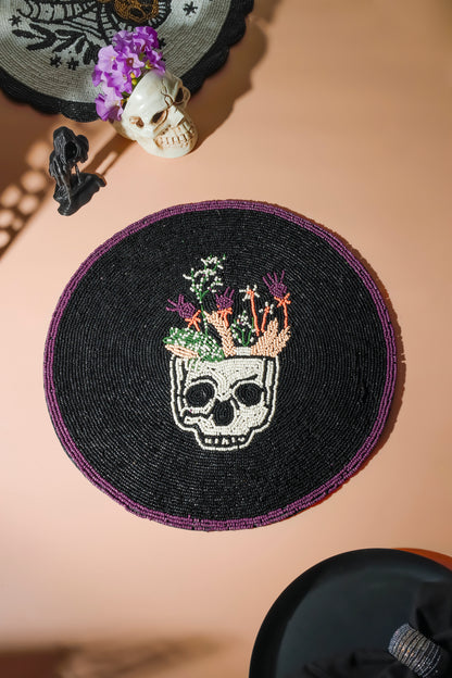 A round black beaded placemat featuring a white skull with colorful flowers growing from its skull cap, styled on a beige surface with gothic props.