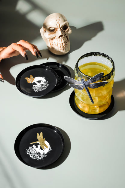 Three black ceramic coasters with gothic designs (skulls and dragonflies) arranged on a light gray surface next to a drink and a decorative skull.